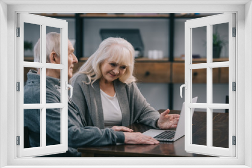 smiling senior couple sitting at table and using laptop at home