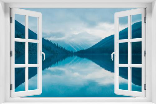 Fototapeta Naklejka Na Ścianę Okno 3D - Amazing silhouettes of mountains and low clouds reflected on mountain lake. Beautiful ripples on water mirror. Cloudy sky in highlands. Atmospheric ghostly landscape. Wonderful mystic mountainscape.