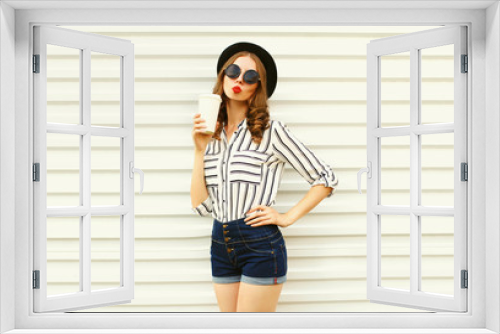 Attractive young woman sending sweet air kiss holding coffee cup in black round hat, shorts, white striped shirt on white wall background