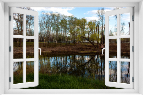 Fototapeta Naklejka Na Ścianę Okno 3D - a small mirror lake surrounded by trees without leaves and a blue sky with clouds floating on it