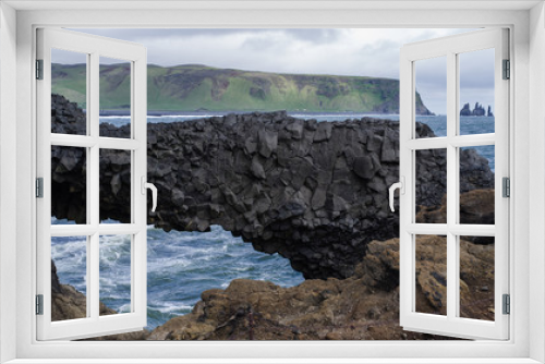 Fototapeta Naklejka Na Ścianę Okno 3D - Summer on Iceland at Dyrholaey nature reserve -a look across a bay with a natural arch made by an eroded volcanic rock.  Ocean waves in centre,  mountains above coast with more sharp cliffs in back