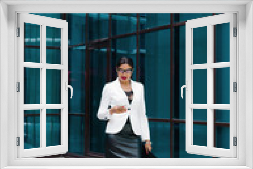 Professional business afro woman in elegant white jacket and skirt with folder with documents in her hands  use smartphone against the background of business center with blue windows