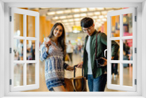 A young and photogenic Asian couple (Korean man, Indian girlfriend in sunglasses) are standing and smiling happily int he middle of an airport during the day. They are about to go for a holiday.