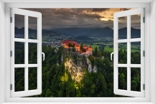 Bled, Slovenia - Aerial drone view of beautiful illuminated Bled Castle (Blejski Grad) with dark rain clouds, golden sunset and the Julian Alps at background at dusk