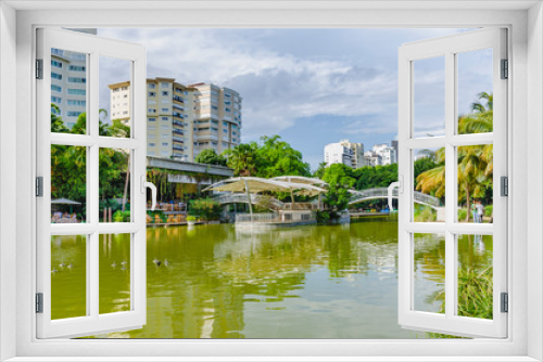 Fototapeta Naklejka Na Ścianę Okno 3D - lake surrounded by natural and tropical landscapes of a park with an arch bridge cruising the lake with beautiful green tones and reflection in the water and city buildings in the background