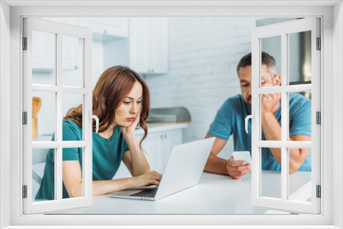 thoughtful man using smartphone while sitting at kitchen table near wife using laptop