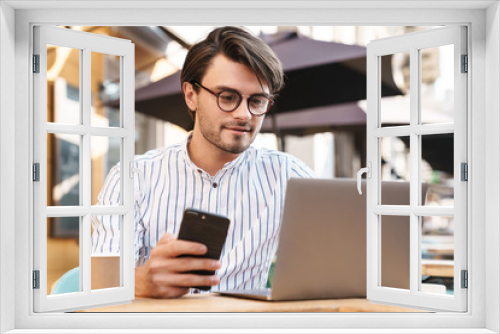 Photo of concentrated caucasian using laptop and cellphone while working in cafe outdoors