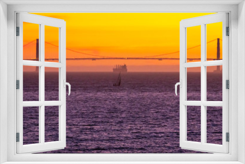Fototapeta Naklejka Na Ścianę Okno 3D - The sun sets over the Golden Gate Bridge as a sailboat and a large container ship set off on the horizon. 