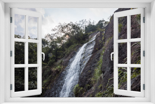 Fototapeta Naklejka Na Ścianę Okno 3D - tall waterfall rushing down the cliff under cloudy sky with rock surface filled with green plants and sunlight shine behind foliage and heavy cloud
