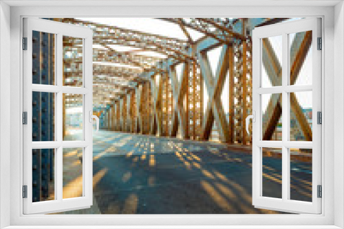 Fototapeta Naklejka Na Ścianę Okno 3D - Asphalt road under the steel construction of a bridge in the city on a sunny day. Evening urban scene with the sunbeam in the tunnel. City life, transport and traffic concept.
