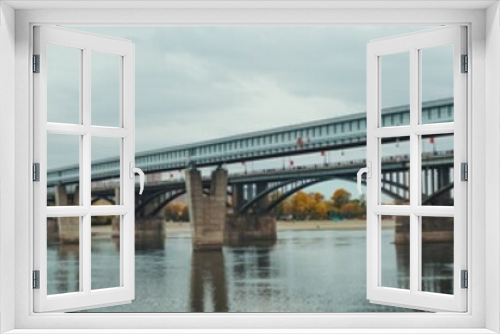 Fototapeta Naklejka Na Ścianę Okno 3D - Panoramic shot of river Ob and its bank in Novosibirsk, Russia during autumn: a metro bridge and transport bridge on the right, the shoreline with yellowed fall trees and cityscape in the background