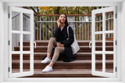 Young pretty casual woman with coffee to go happily talking on cellphone in city park