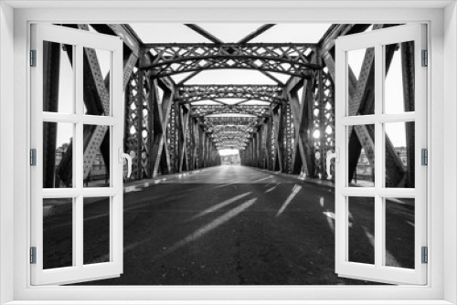 Fototapeta Naklejka Na Ścianę Okno 3D - Black and white asphalt road under the steel construction of a bridge in the city on a sunny day. Evening urban scene with the sunbeam in the tunnel. City life, transport and traffic concept.	
