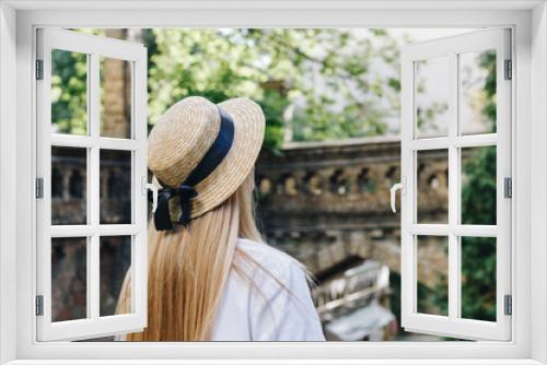 Fototapeta Naklejka Na Ścianę Okno 3D - A thin blonde girl with long hair in a white dress stands with her back to the camera and looks into the distance in a wide-brimmed hat. City landscape, traveler.