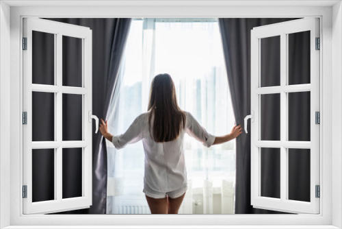 Beautiful young woman opening curtains and looking through the window
