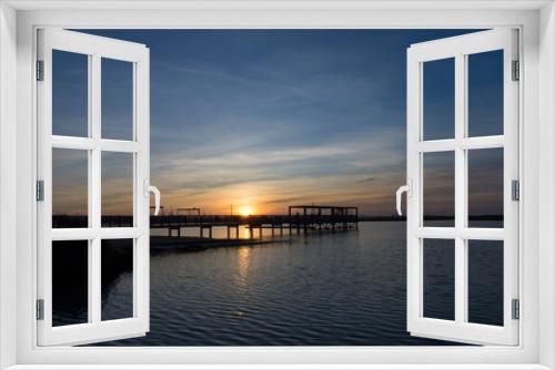 Fototapeta Naklejka Na Ścianę Okno 3D - coastal landscape with wooden jetty reflected in calm waters under a sunset with clouds and large industrial cranes in the background
