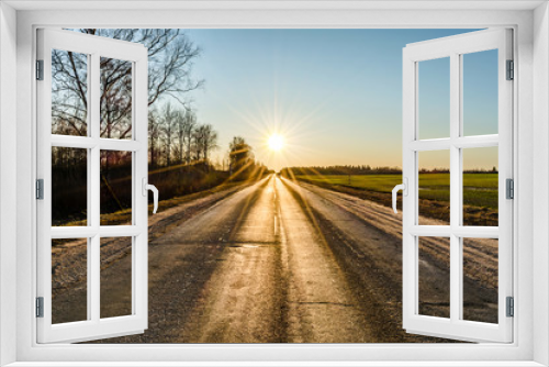 Fototapeta Naklejka Na Ścianę Okno 3D - The asphalt of an old road, cracked in the cold, is lit by the sun's rays in a haze of blue sky against the background of a overed fields and trees