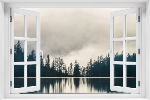 Fototapeta Naklejka Na Ścianę Okno 3D - Silhouettes of pointy fir tops on hillside along mountain lake in dense fog. Reflection of coniferous trees in shiny calm water. Alpine tranquil landscape at early morning. Ghostly atmospheric scenery