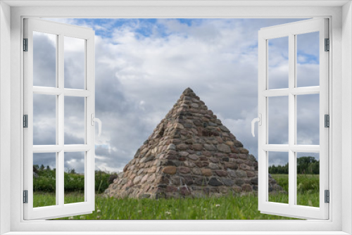 Fototapeta Naklejka Na Ścianę Okno 3D - Stone pyramid, made of large granite stones. Green grass and trees, blue sky, storm clouds. Natural environment concept and building with four walls, Latvia, Europe