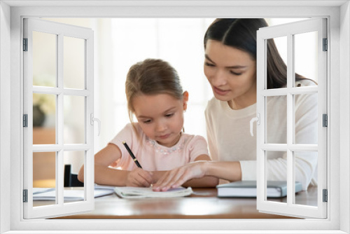 Head shot focused attentive little girl studying, doing homework with mother. Affectionate young mother teaching small daughter reading writing. Female teacher giving private lessons to kid at home.