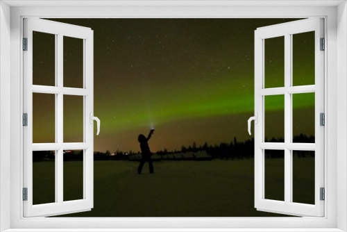 Fototapeta Naklejka Na Ścianę Okno 3D - Woman with flashlight before green aurora borealis at night in snow landscape, green and orange sky, cottages in background, Lapland, Finland