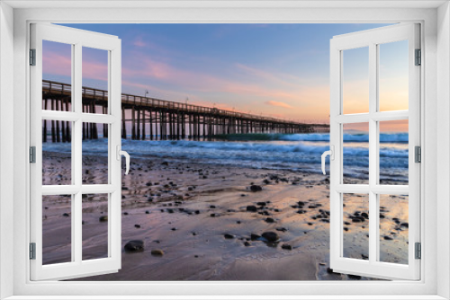 Fototapeta Naklejka Na Ścianę Okno 3D - Ventura Pier at sunset, Ventura, California. Rocks and sand in foreground, water receded in low tide, with incoming waves. Lamps on pier; colored twilight sky in background.