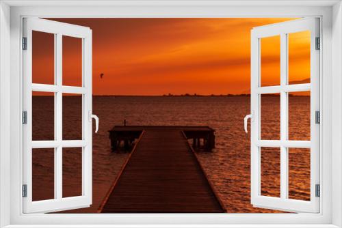 Fototapeta Naklejka Na Ścianę Okno 3D - Wooden pier on a warm light during sunset, with orange sky and a Kite surf and two mountains silhouette in the background