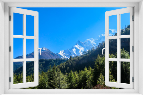 Fototapeta Naklejka Na Ścianę Okno 3D - A view on Himalayan valley along Annapurna Circuit Trek, Nepal. There is a dense forest in front. High, snow caped mountains' peaks catching the sunbeams. Serenity and calmness. Barren slopes