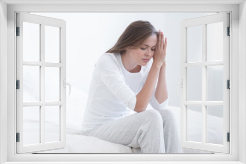 Young female sitting on white bed, pressing palms to one side of face, as if she can not endure daylight, suffering from insomnia or migraine