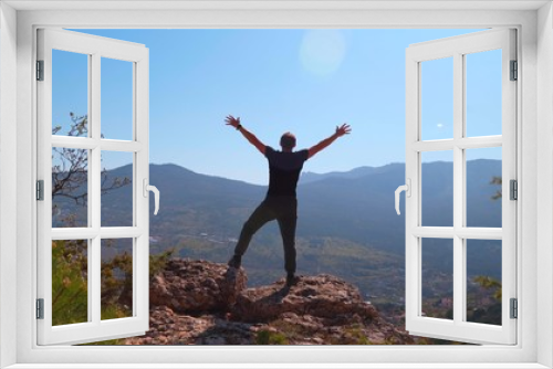Fototapeta Naklejka Na Ścianę Okno 3D - Young inspired man raises his hands up standing on the top of a mountain and looks at a beautiful valley of forest against beautiful blue sky. Silhouette of a happy man standing.