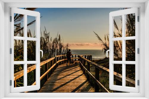 Fototapeta Naklejka Na Ścianę Okno 3D - person with his dog who goes to the beach by a wooden bridge in a beautiful, warm and colorful sunrise