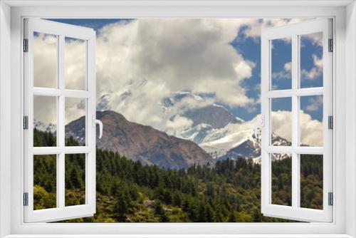 Fototapeta Naklejka Na Ścianę Okno 3D - Clouds cover the snow covered peak of Nilgiri North towering above a green, alpine forest in the Himalayan village of Tukuche on the Annapurna Circuit in Nepal.