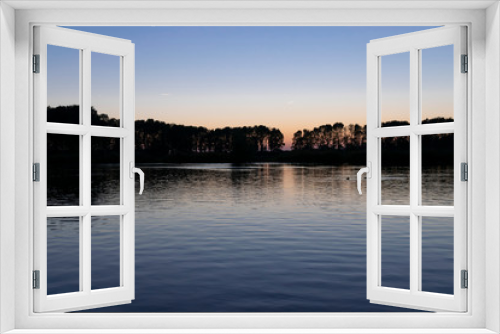 Fototapeta Naklejka Na Ścianę Okno 3D - The surface of the water with ripples and reflections of sunset on background of a calm symmetrical landscape in a light misty haze, soft focus.