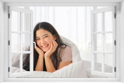 Happy Asian girl with black long hair lying on the bed with hands touching her face and giving sweet smile and giving eye contact with copy space