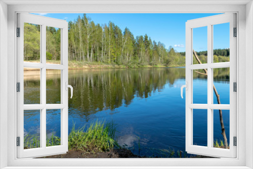 Fototapeta Naklejka Na Ścianę Okno 3D - spring landscape with a river, in the waters of which clouds are reflected, the banks of the river are covered with trees, the first spring greenery in nature, stones and green grass in the foreground