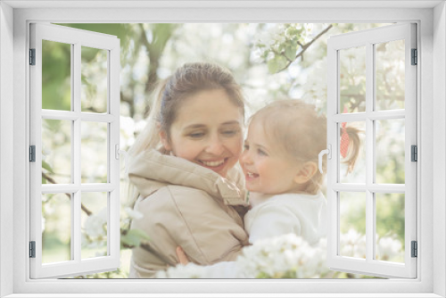 A beautiful charming mother and daughter walk in a flowering park in spring. Warm hugs, tenderness, kiss mom and baby. Blooming white trees, the sun, good mood, joy.