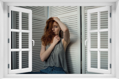 Redhead girl with freckles in a T-shirt sits leaning on a white wooden screen, and looks into the frame, covering face with hair.