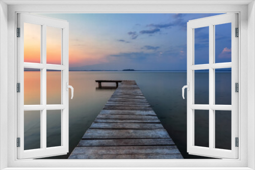 Fototapeta Naklejka Na Ścianę Okno 3D - Old wooden jetty, pier reveals views of the beautiful lake, blue sky with cloud. Sunrise enlightens the horizon with orange warm colors. Summer landscape. Free space for text.
