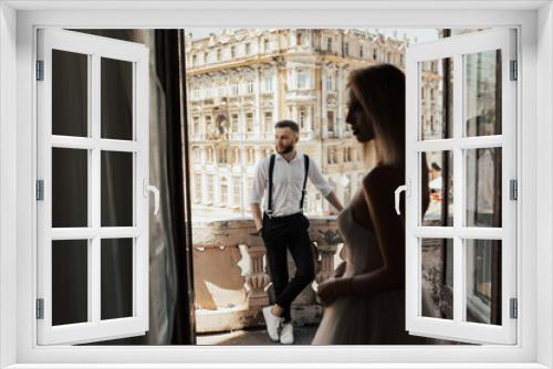 Wedding couple stand on a hotel balcony with beautiful architecture on background, view through an open antique window. Newlyweds on a terrace, balcony. Happy wedding day of marriage.