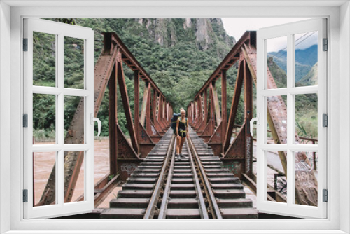 Fototapeta Naklejka Na Ścianę Okno 3D - Portrait of positive young woman with tourist backpack smiling at camera and standing on rails of bridge while travelling in wildness nature with high mountains and beautiful scenery on background
