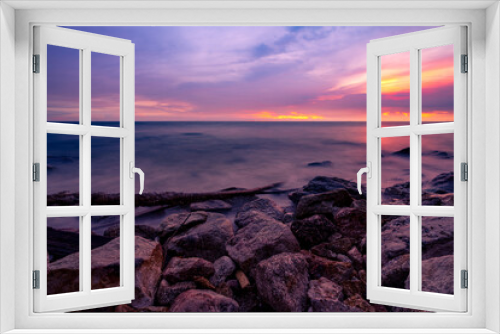 Fototapeta Naklejka Na Ścianę Okno 3D - Stone beach and calm sea at dusk with purple sunset sky. Tropical sea. Skyline in the evening with golden, purple, and red sky. Seascape. Summer vacation travel. Rock coast. Beauty in ocean nature.