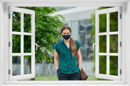 Sad woman in a medical face mask walks between trees thrusting hands into pockets of trousers in downtown. Girl keeping social distance wears a protective face mask to avoid the spread of coronavirus