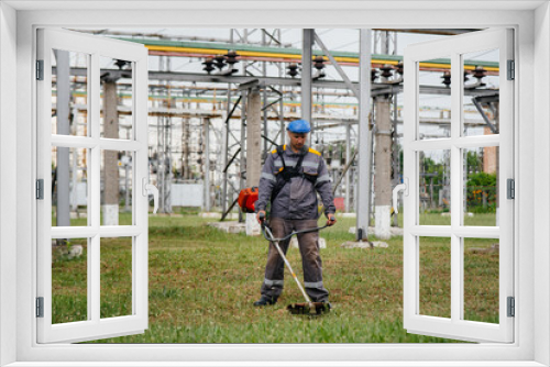 Fototapeta Naklejka Na Ścianę Okno 3D - A young man mowing grass on the territory of an electric substation in overalls. Grass cleaning at the enterprise, implementation of fire safety measures