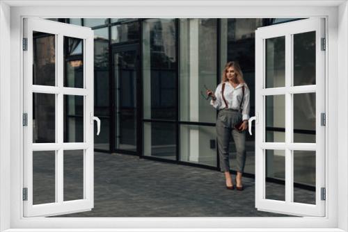 Full length portrait of young female CEO, manager, director, glass skyscraper. Business concept. Feminine gait. Gorgeous agent or entrepreneur holding sunglasses and clutch bag. Copy space.