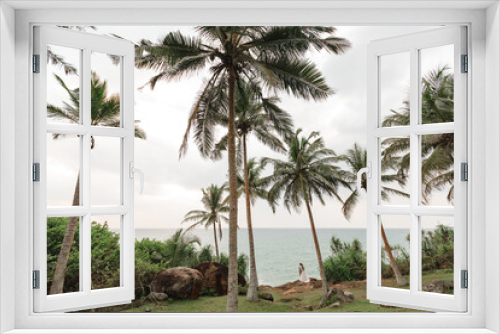 Fototapeta Naklejka Na Ścianę Okno 3D - Just married couple standing on sea cliff under palm tree and enjoy view on tropical sand beach. Back view bride and groom looking at the blue ocean