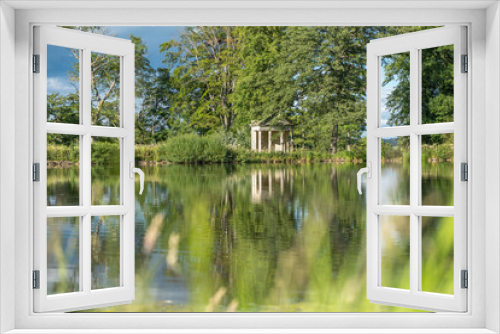 Fototapeta Naklejka Na Ścianę Okno 3D - A view with blurred grass in the foreground across a beautiful lake with reflections of trees towards an old abandoned structure in the middle of a rural countryside scene