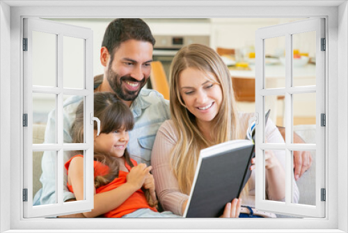 Joyful parents couple and little black haired girl sitting on couch in living room, reading book together and laughing. Childhood or literature concept