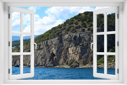 Fototapeta Naklejka Na Ścianę Okno 3D - Seascape from the sea. A rocky coast that flows into the sea. Coast against the background of mountains and greenery. Clouds over the mountains