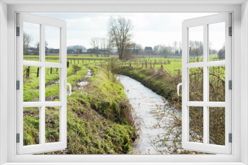 Fototapeta Naklejka Na Ścianę Okno 3D - A little swollen river, turns left. The water is brownish,  grassy banks on each sides, meadows, fields, houses in the background.