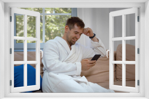 Attractive smiling young man wearing bathrobe sitting on couch at living room using mobile phone